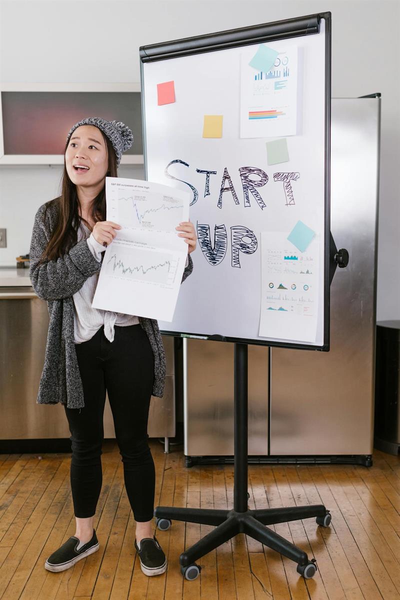 Junge Frau, die vor einem Whiteboard steht, auf dem Start-up geschrieben steht, hält ein Blatt Papier mit Aktiencharts in der Hand.