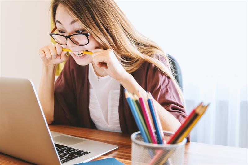 Gestresste Frau mit Brille am Laptop beisst in gelben Bleistift