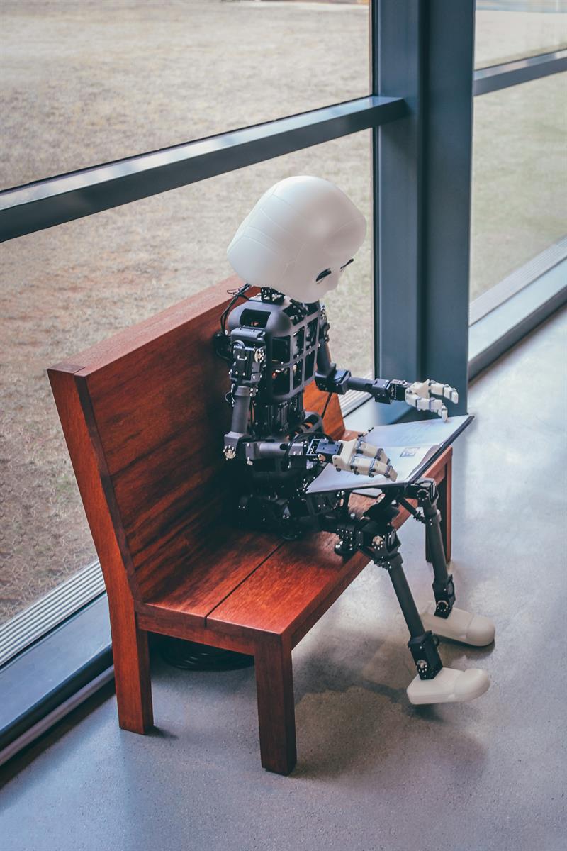 Ein Roboter sitzt vor einer Glasfront auf einer Holzbank und liest ein Buch.