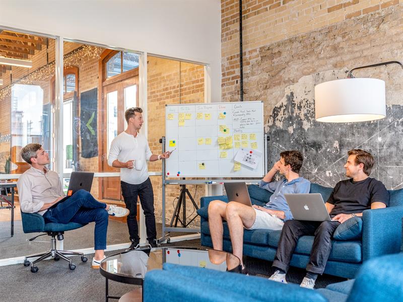 Drei Männer mit Laptops sitzen um Flipchart, vor dem ein Mann steht und erklärt