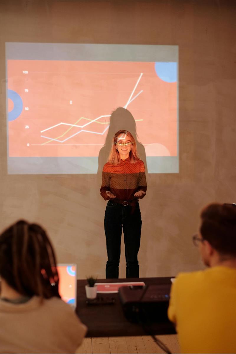 Junge Frau, die vor einer Leinwand steht und eine Präsentation hält. Die Leinwand zeigt ein Diagramm mit mehreren Linien, die einen positiven Aufwärtstrend darstellen.