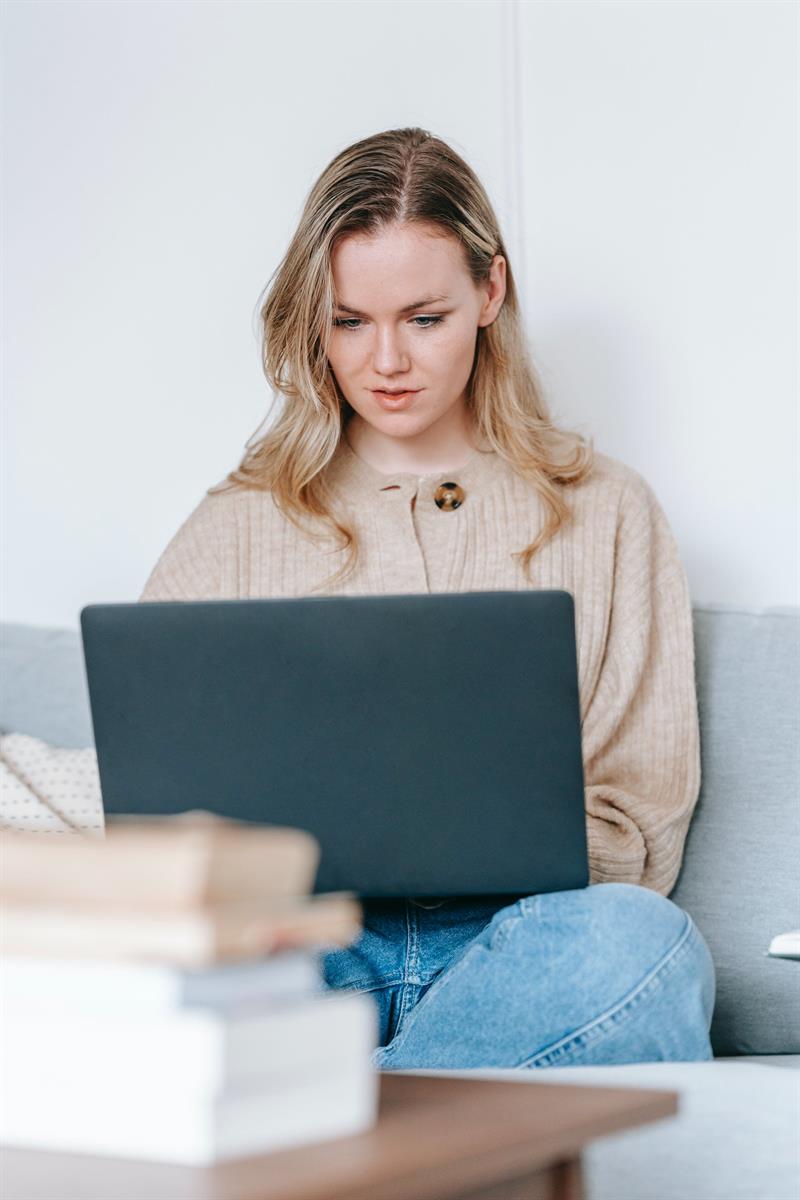 Junge Frau mit Laptop auf dem Schoß auf Sofa und Büchern auf dem Tisch vor ihr